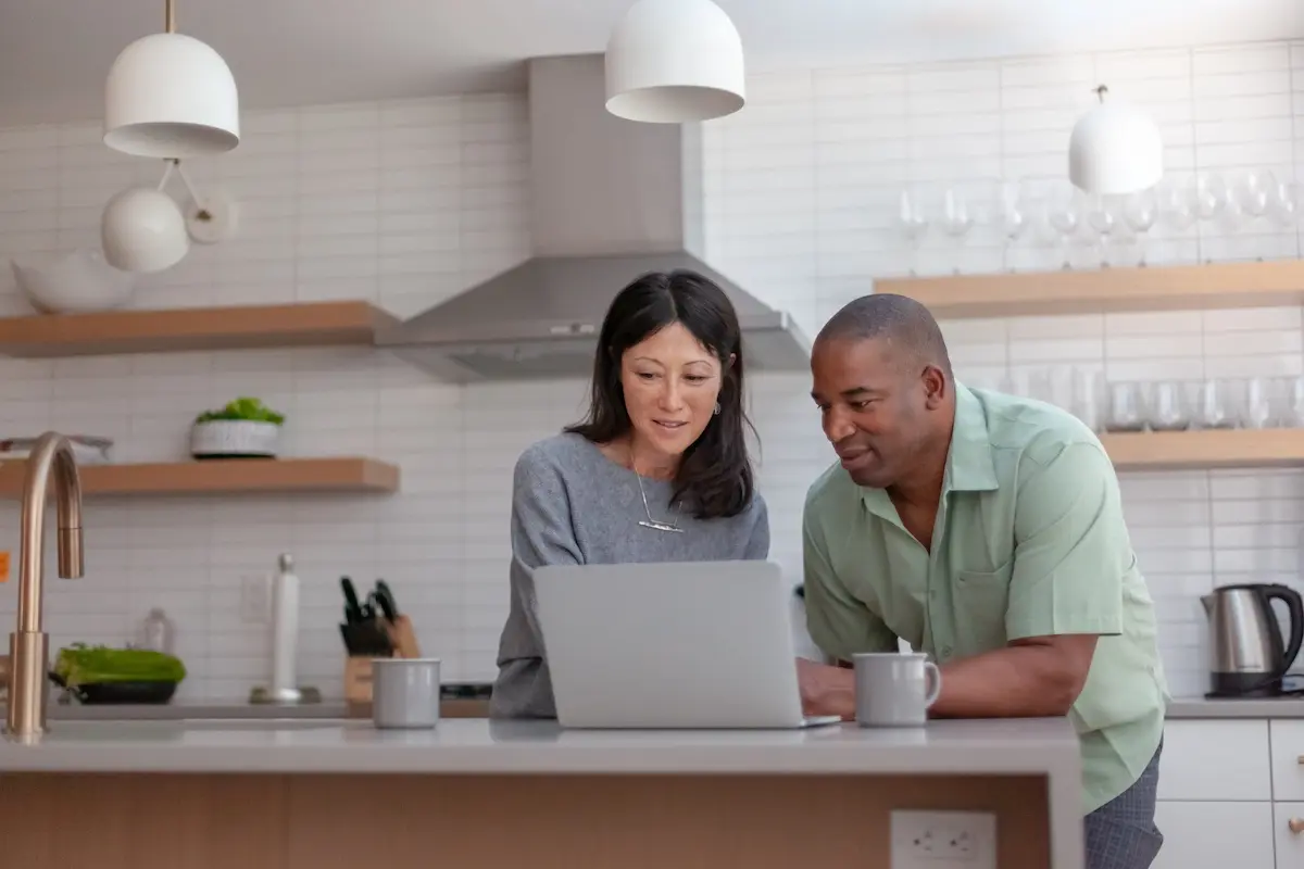 A couple standing in a modern kitchen reviewing information together on a laptop at the counter.